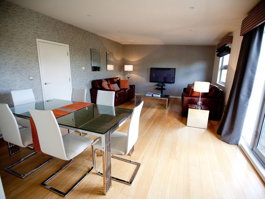 Una sala de estar moderna con una mesa de comedor de vidrio, sillas blancas, un sofá de cuero, un televisor de pantalla plana y un amplio ventanal con cortinas.