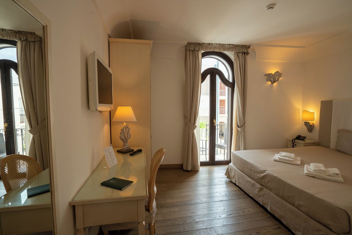 A hotel room with a bed, a desk with a lamp, a chair, a mirror, and a view through a window with curtains.
