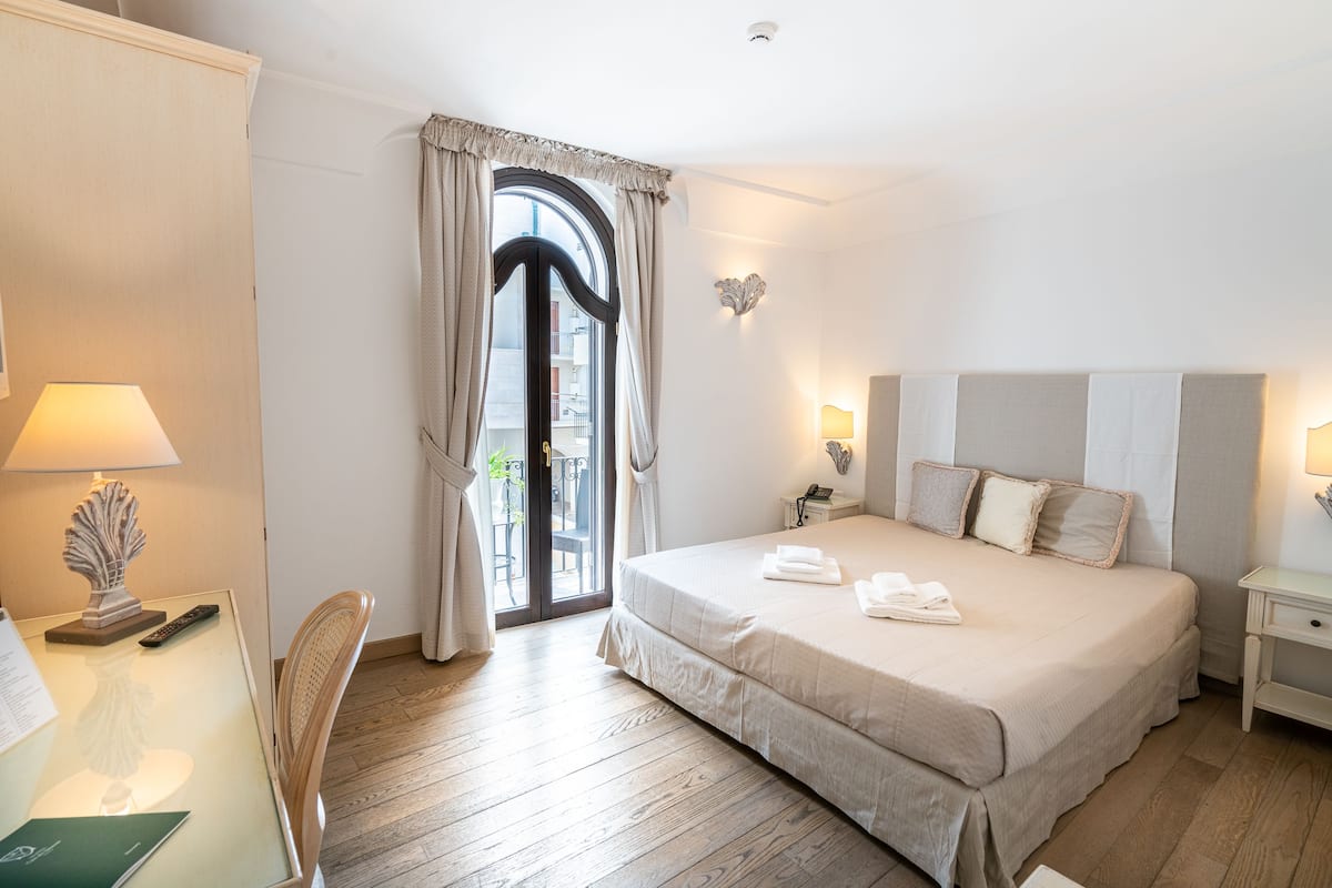 Habitación de hotel con una cama grande, un escritorio con lámpara, una silla y vistas a través de una ventana con cortinas.
