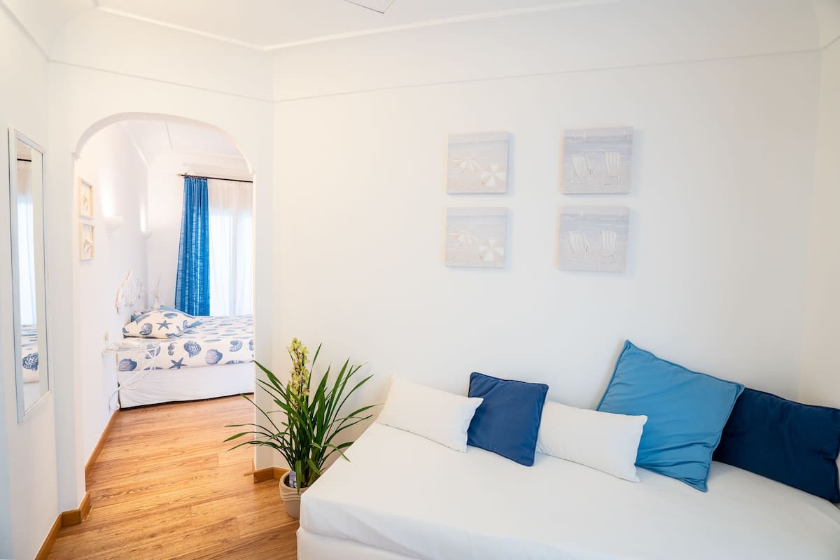 Un dormitorio moderno con una cama blanca, almohadas azules y suelo de madera. Hay una planta en el suelo y una ventana con cortinas azules.