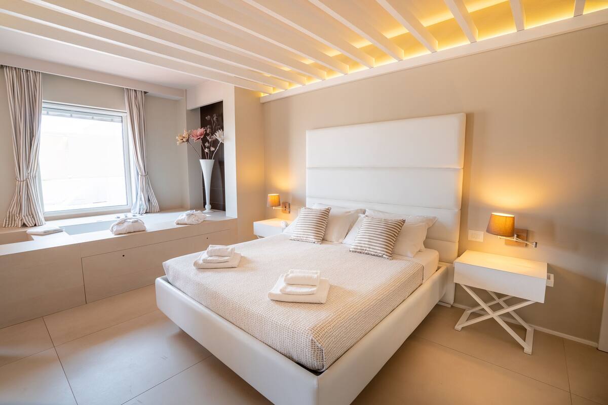 Un dormitorio moderno con una cama grande, lámparas de noche, una ventana con cortinas y una mesita de noche con un florero.