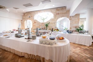 Una mesa larga con diversos platos y alimentos, dispuesta para un bufé, frente a una pared de piedra con ventanas arqueadas y una escalera.