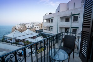 Un balcón con una mesa de vidrio y una silla de mimbre, con vista a un pueblo costero con edificios y al mar.