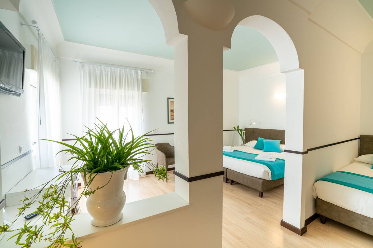 Habitación de hotel moderna con una planta en maceta grande, cama con ropa de cama color turquesa y vista a otra habitación a través de un arco.