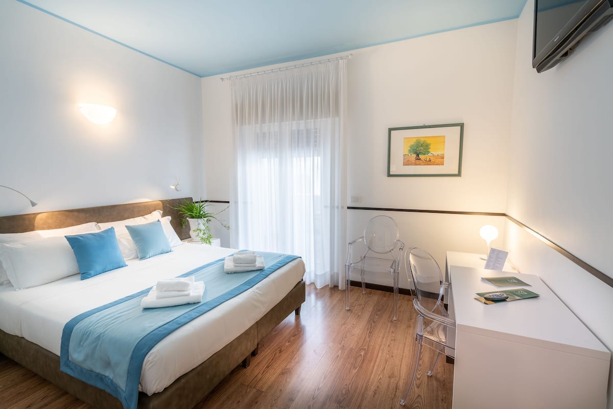 Una habitación de hotel moderna con una cama bien hecha, un escritorio con lámpara y un ventanal con cortinas translúcidas.