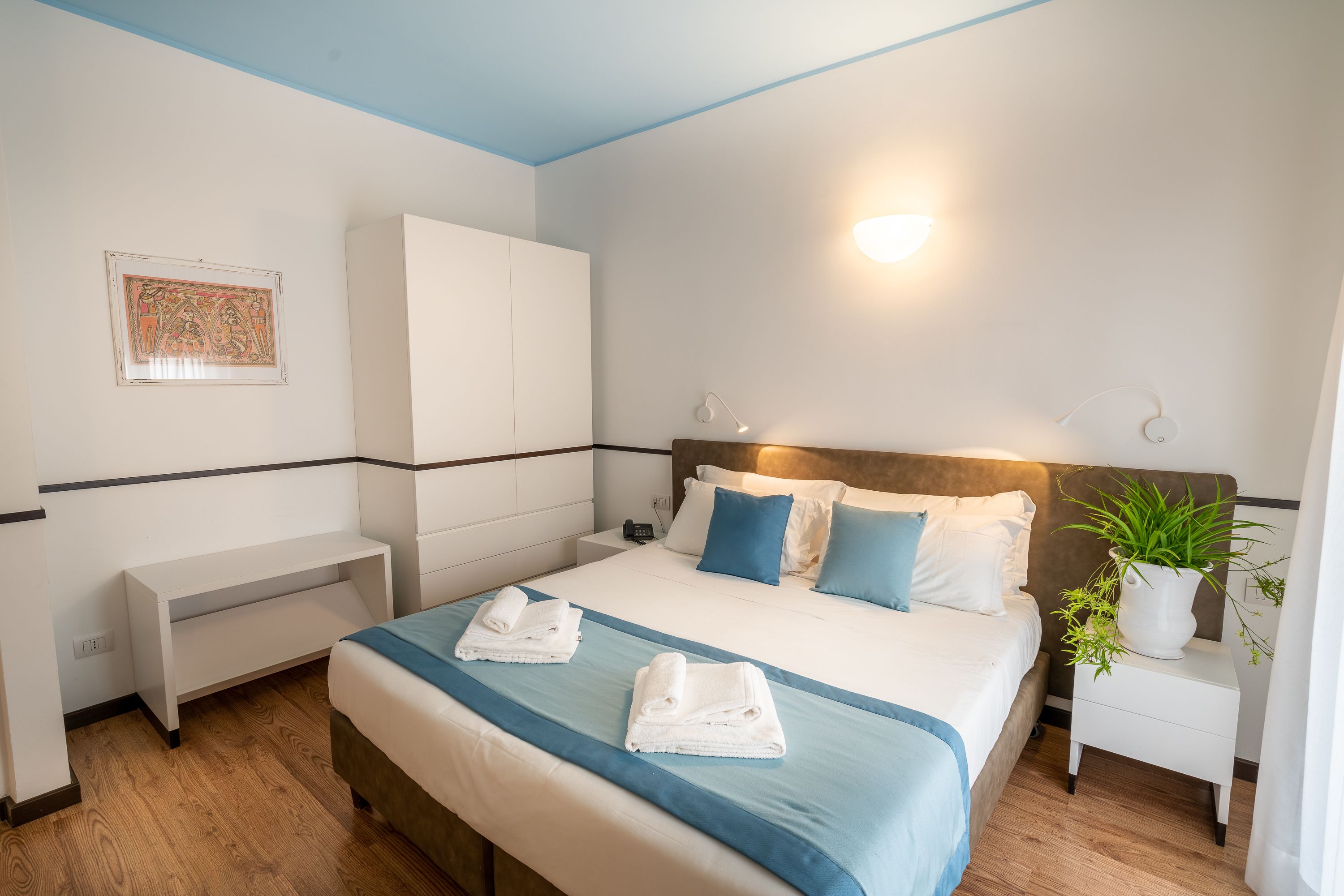 Habitación de hotel con cama, mesita de noche y una planta en maceta.