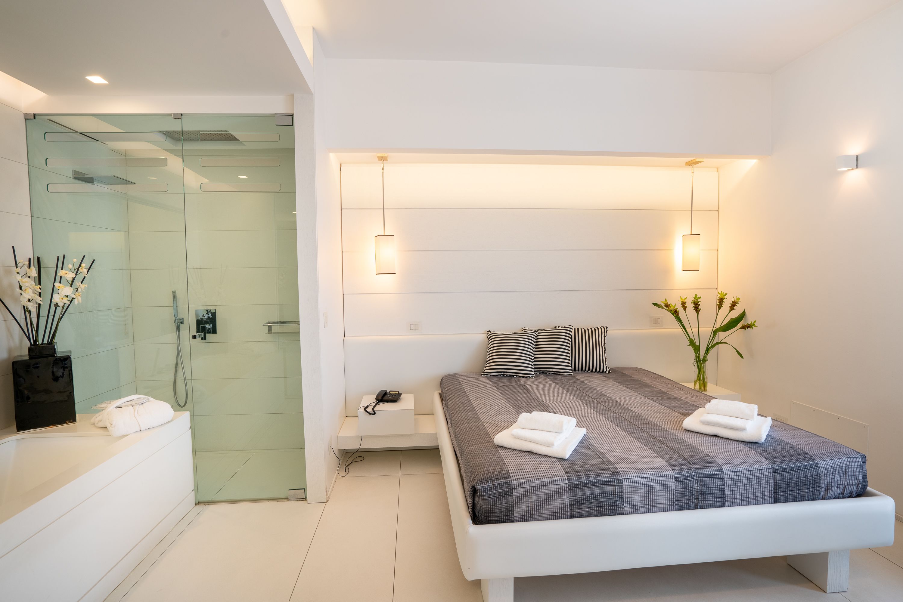 Een moderne slaapkamer met een groot bed, nachtkastjes, een douchecabine van glas en een klein nachtkastje met een telefoon en oplader.