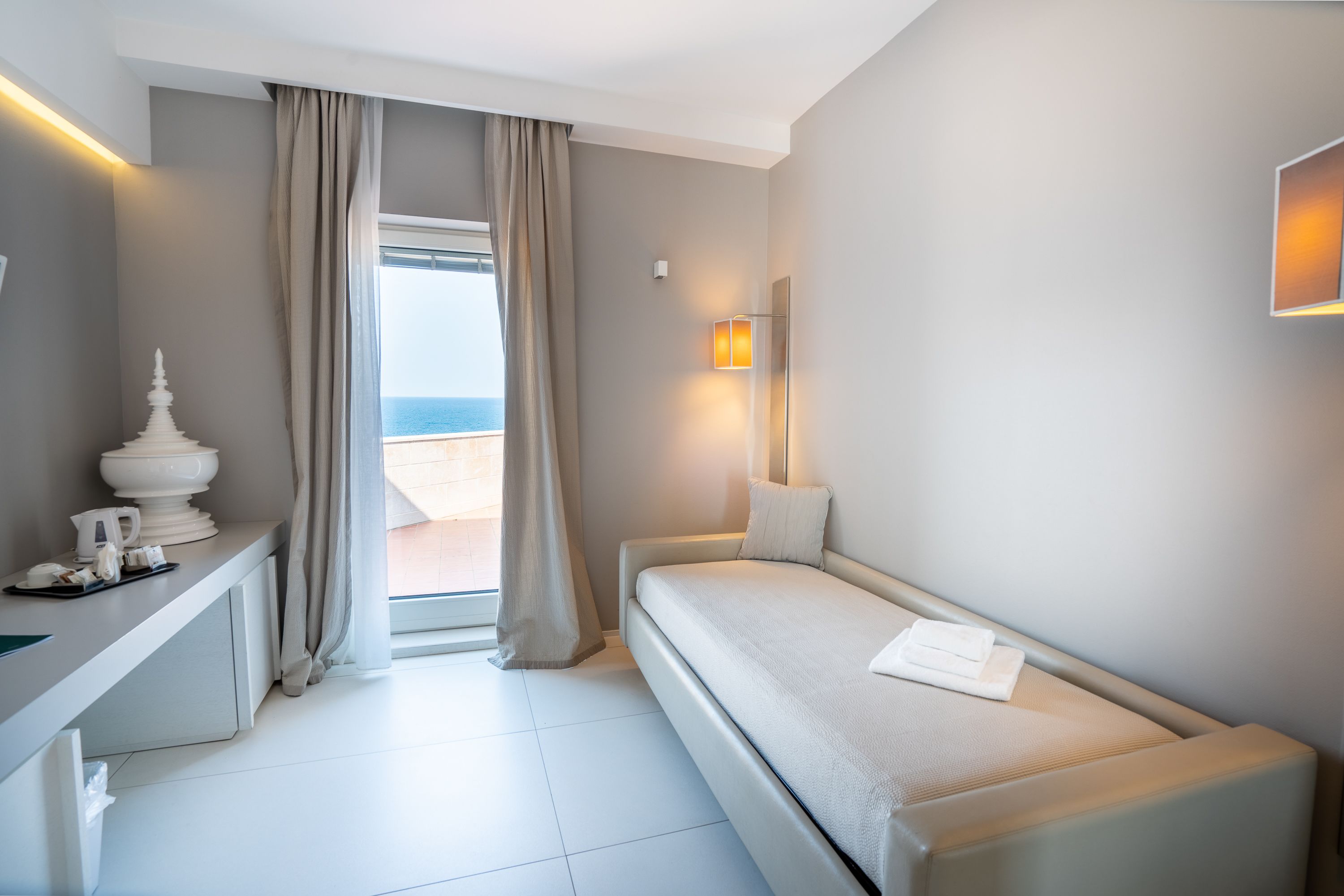 A modern hotel room with a bed, a desk, and a view of the sea.