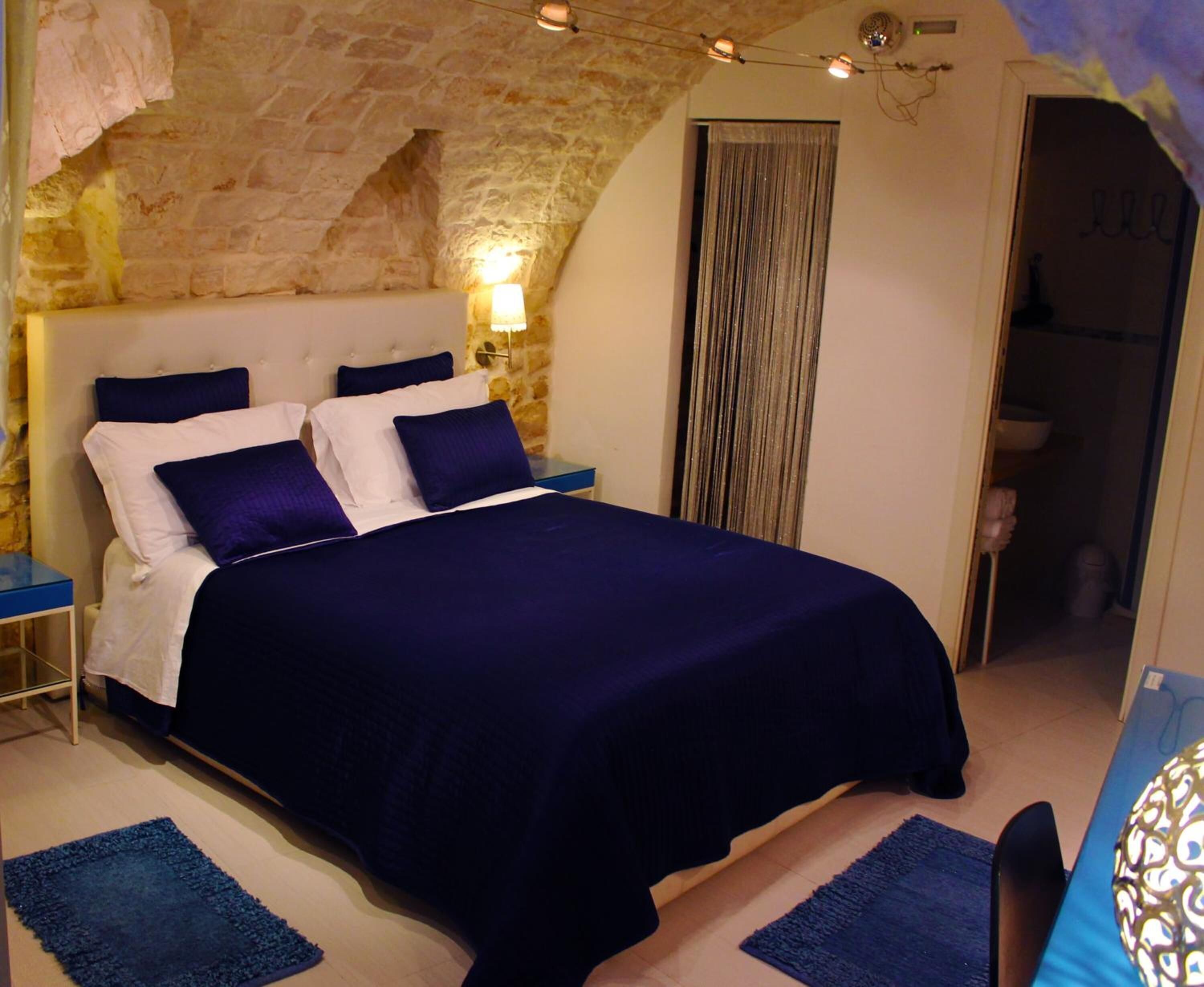 A bedroom with a stone wall, a bed with a dark blue bedspread, two bedside tables, and a wall-mounted lamp.