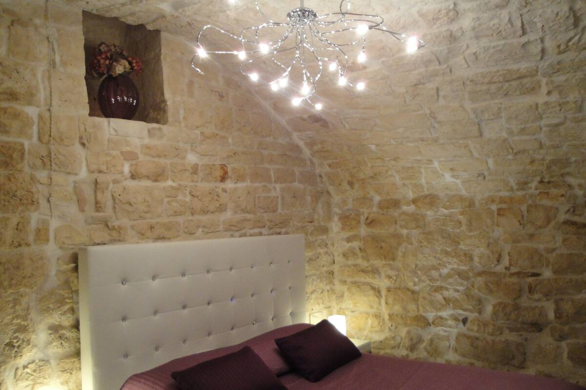 A bedroom with a stone wall, a white tufted headboard, and a bed with a red bedspread and two dark pillows.