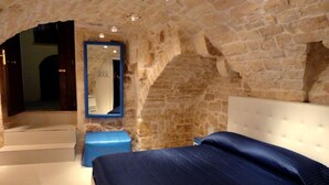 A bedroom with a stone wall, a large mirror, a bed, and a small blue ottoman.