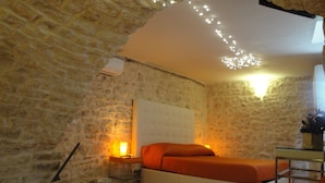 A room with stone walls, a bed with an orange bedspread, a bedside table with a lamp, and a small table with a plant.