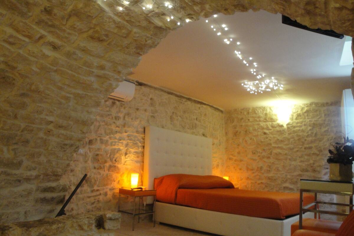 A room with stone walls, a bed with an orange bedspread, a bedside table with a lamp, and a small table with a plant.