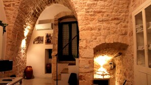 A narrow stone corridor with an arched doorway leading to a room with a bed and a window.