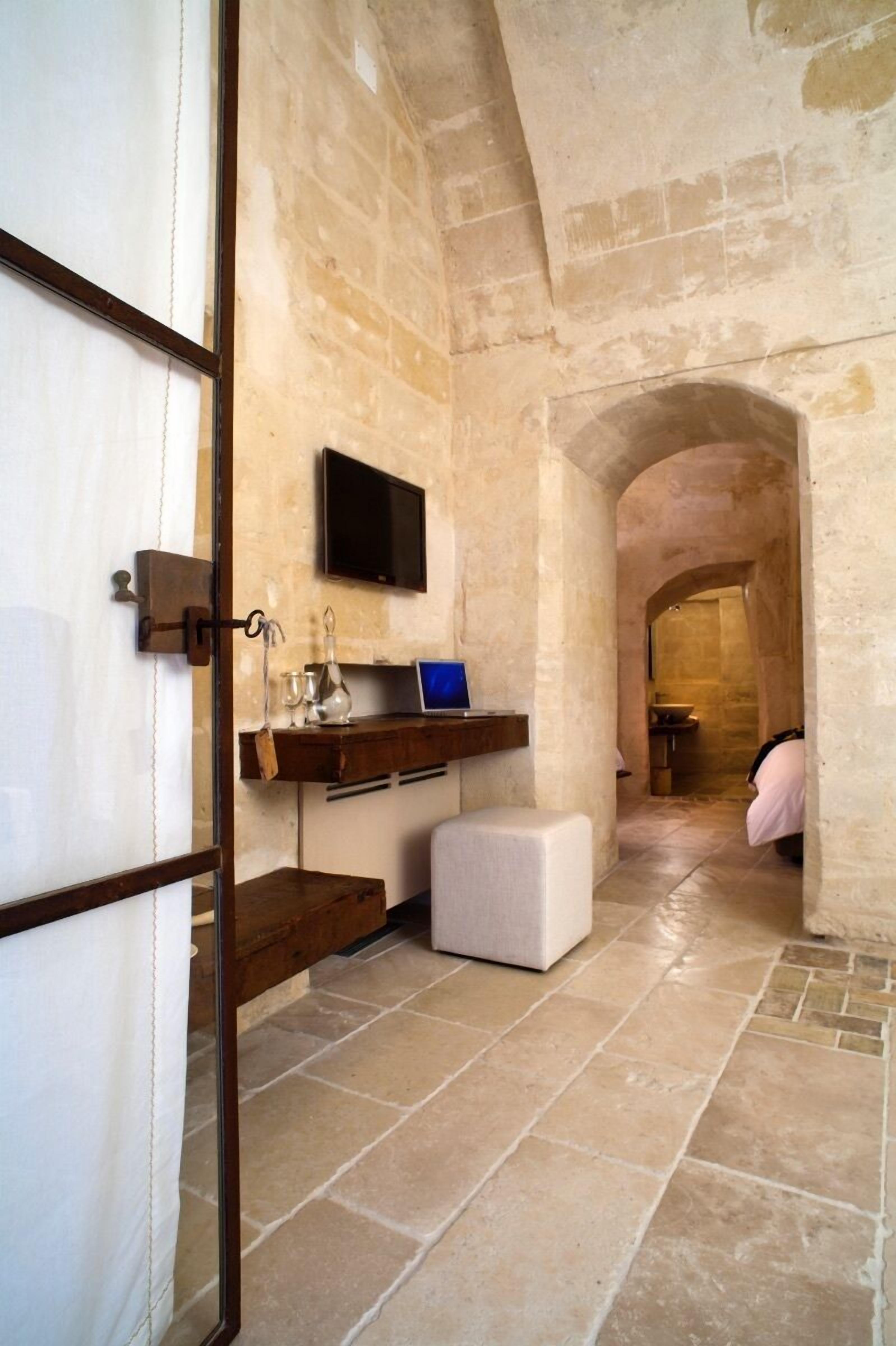 A narrow stone-walled hallway with a television mounted on the wall, a small bench, and a doorway leading to another room.