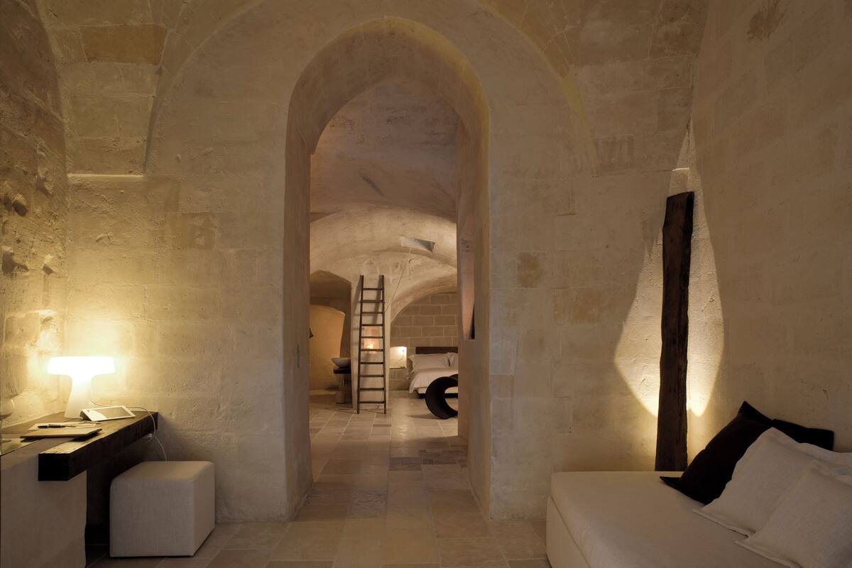 A narrow passageway with stone walls and a wooden ladder leading to a bedroom with a bed and a nightstand.
