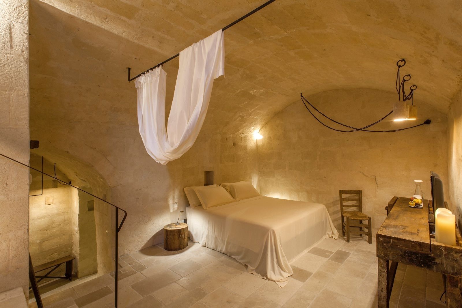 Une chambre à coucher avec un lit, une chaise en bois, une petite table avec une bougie et un luminaire suspendu.