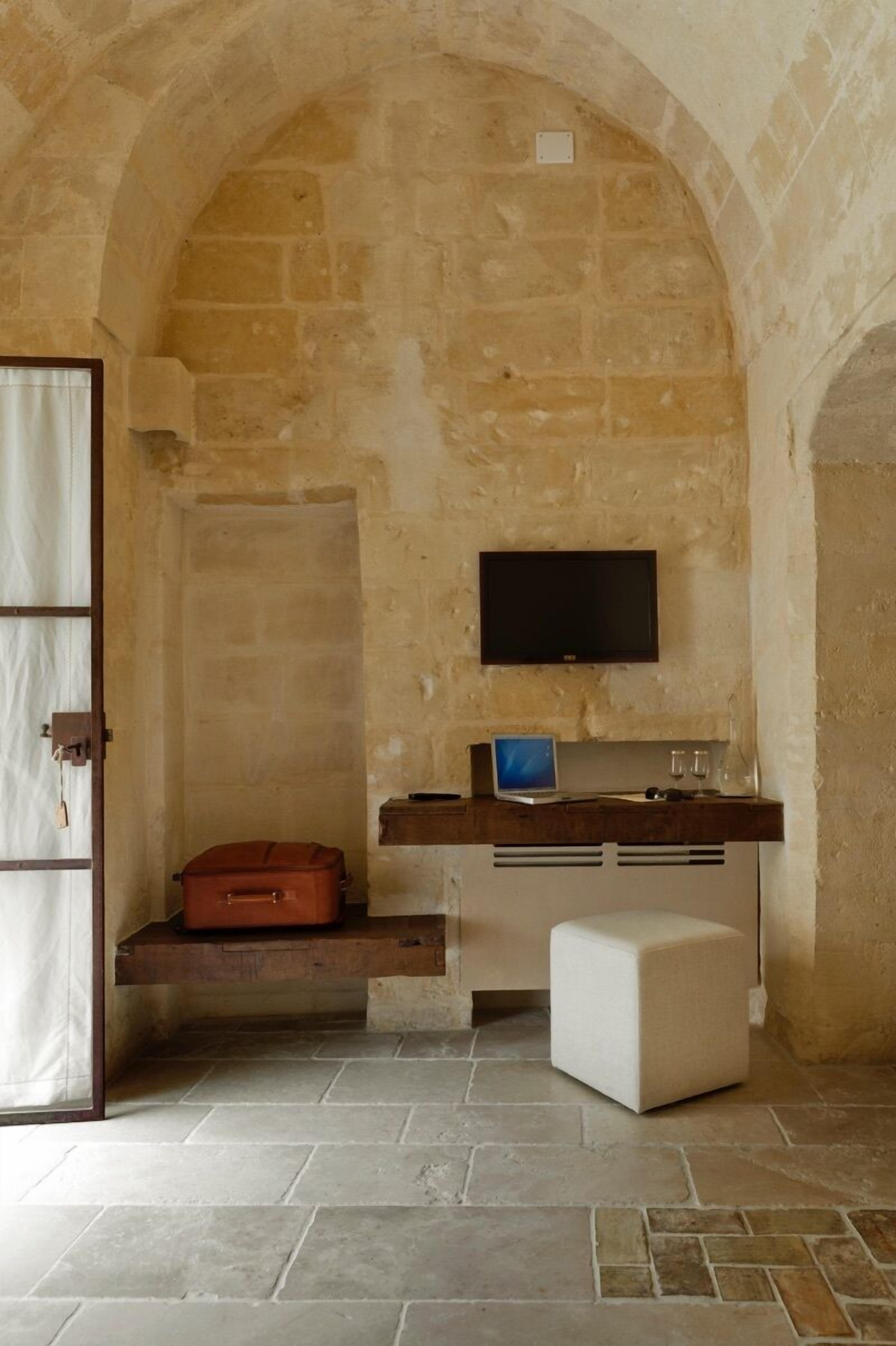 Una habitación con paredes de piedra, un televisor montado, una maleta, una computadora portátil y un pequeño otoman.