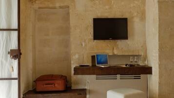 Una habitación con paredes de piedra, un televisor montado, una maleta, una computadora portátil y un pequeño otoman.