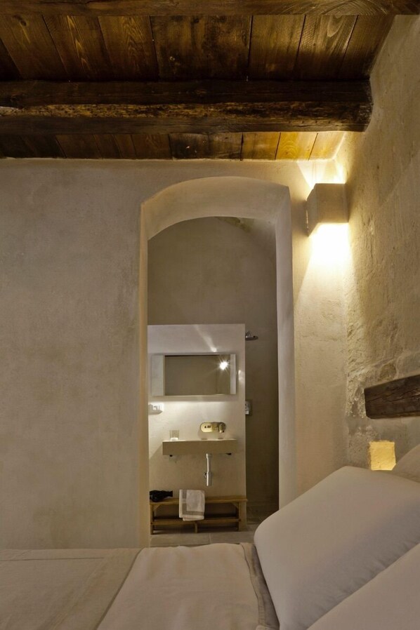 A cozy bedroom with a wooden ceiling, a white bed, a small bedside table, and a built-in bench. - Corte San Pietro (Matera)