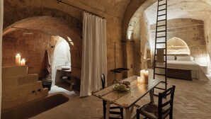 A rustic stone room with a wooden table, a bench, a ladder, a bed, and a bathtub. - Corte San Pietro (Matera)