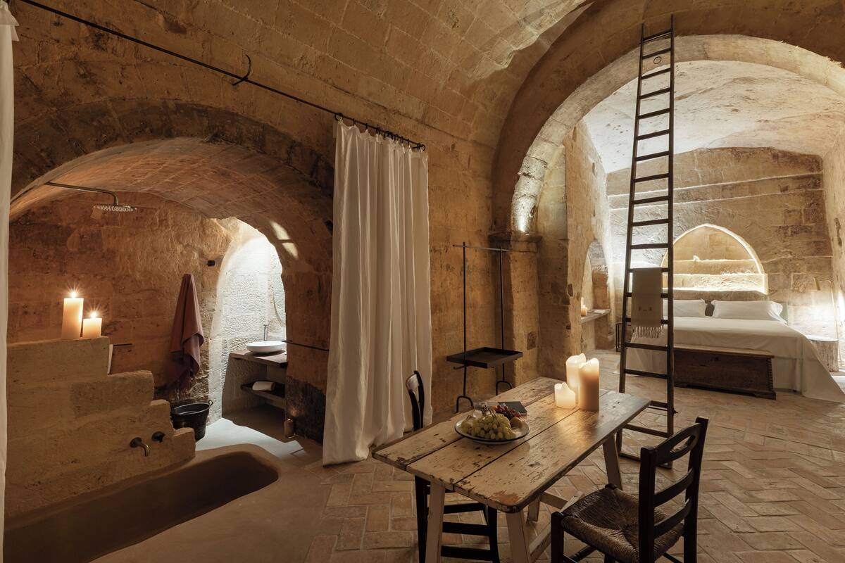 A rustic stone room with a wooden table, a bench, a ladder, a bed, and a bathtub.