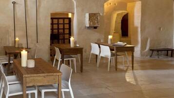 A rustic dining area with wooden tables and white chairs, featuring stone walls and a tiled floor.