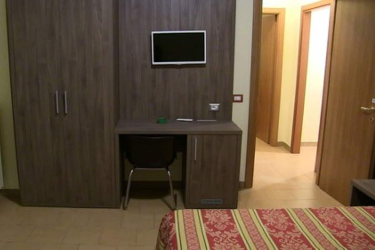 Un dormitorio con un armario de madera, un escritorio con silla, una cama con colcha estampada y un televisor montado en la pared.