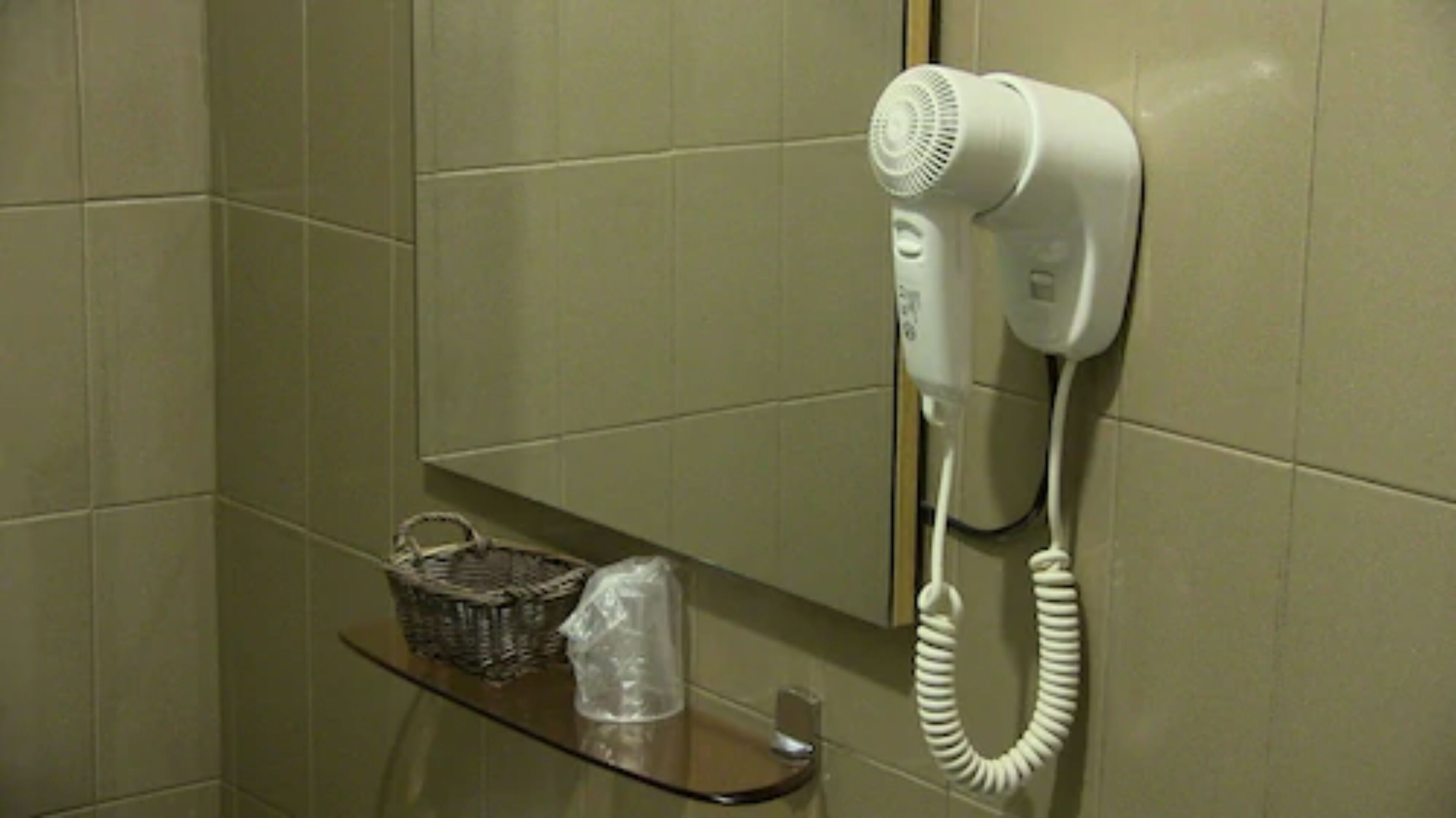 A hairdryer mounted on a wall in a bathroom with a basket and a plastic container on a shelf.