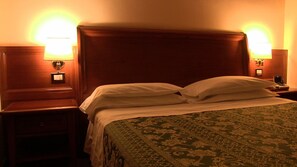 Una habitación de hotel con cabecera de madera, dos lámparas de noche, una mesita de noche y una colcha estampada.