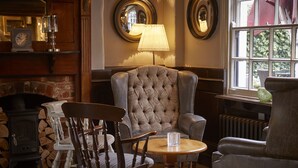 A cozy room with a fireplace, a large armchair, a wooden table, and a window with a view of greenery. - Two Brewers by Chef and Brewer Collection (Kings Langley)