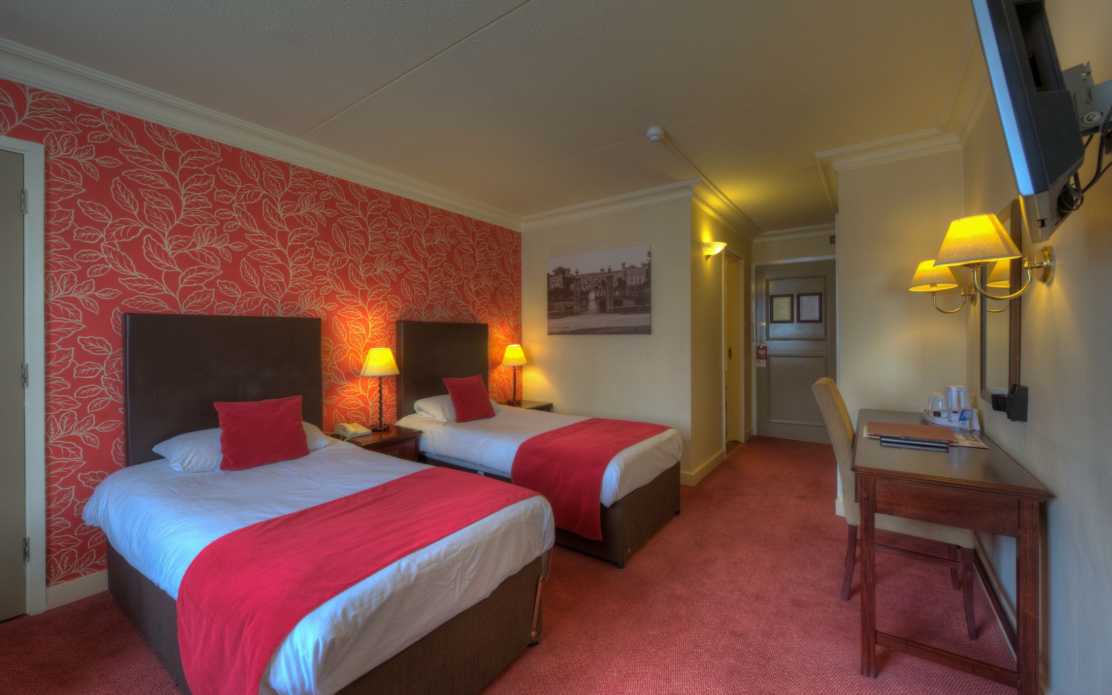 A hotel room with two beds, red and white bedding, a desk with a chair, and a television on the wall.