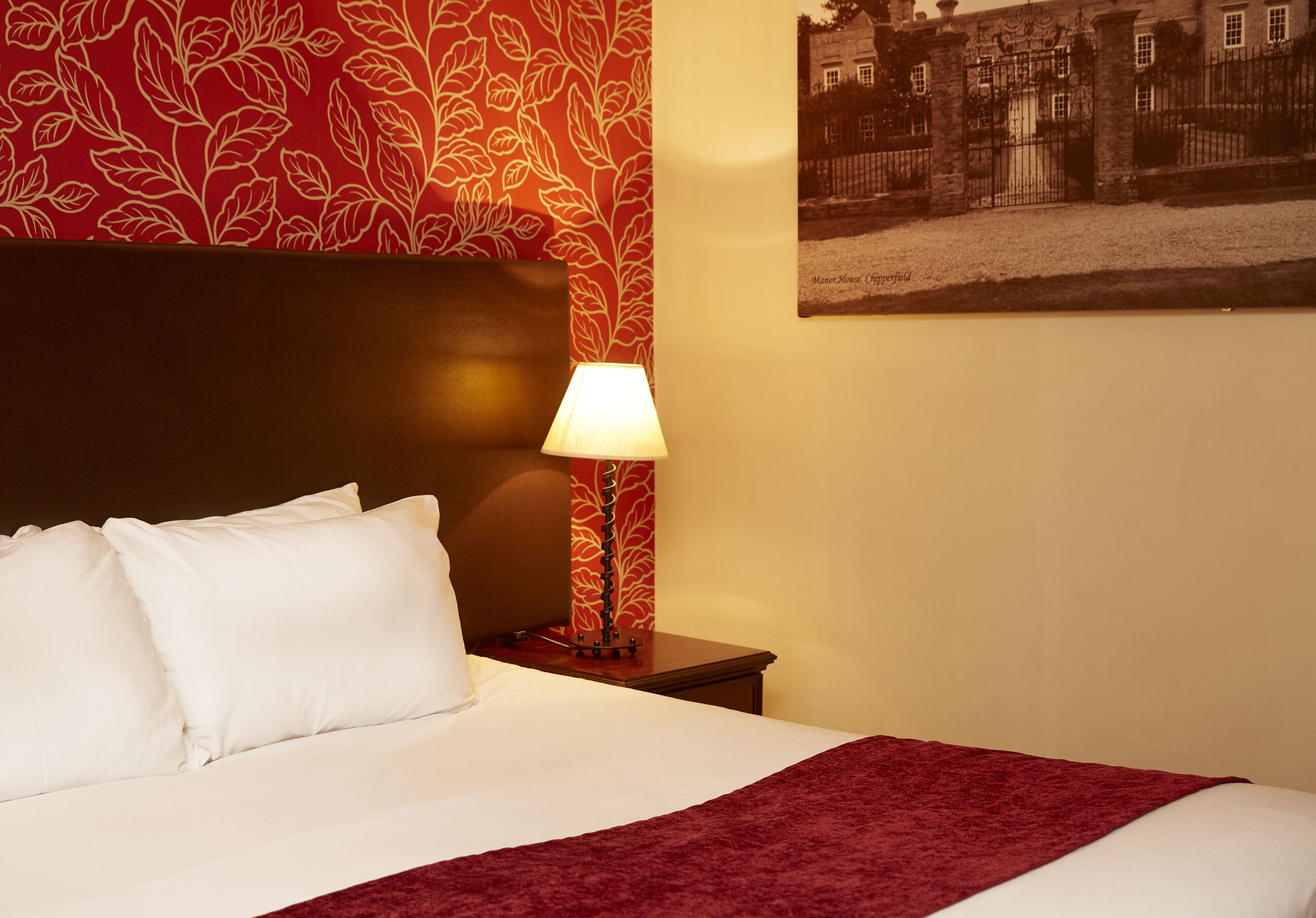 A hotel room with a bed, white pillows, a red blanket, a lamp, a nightstand, and a wall with a floral pattern and a framed picture.