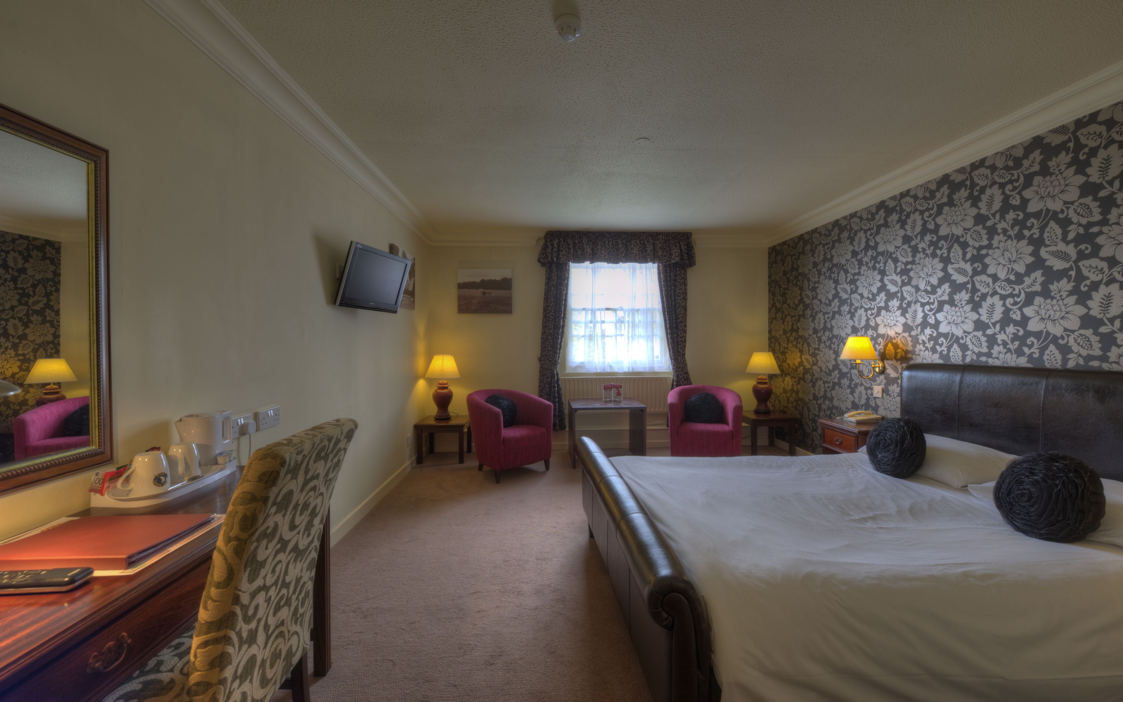 Habitación de hotel con cama, dos sillas, escritorio con espejo, televisor y ventana con cortinas.