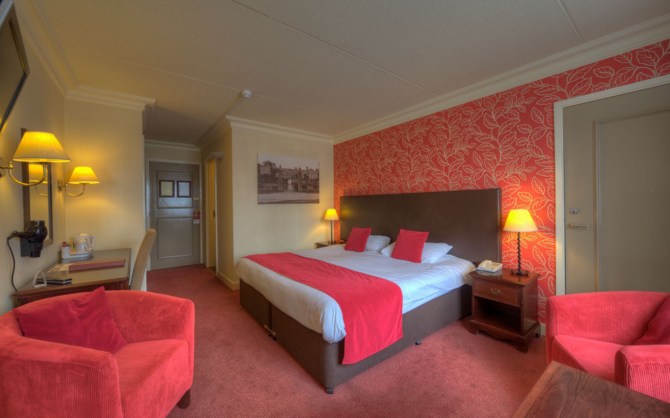 A hotel room with a large bed, two bedside tables with lamps, a chair, and a red carpeted floor.