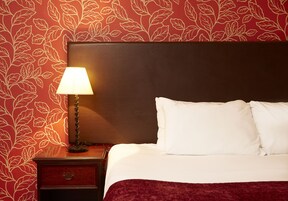 Una cama bien tendida con almohadas blancas y un cabecero oscuro, una lámpara de noche y una mesita de noche de madera con un cajón.