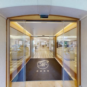 A hotel entrance with a checkered floor, a logo on the mat, and glass doors leading to a lobby with seating and tables. - Relais La Fattoria (Melfi)