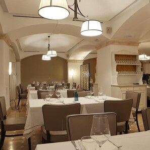 A dining area with multiple tables set for service, overhead lighting, and a kitchen area in the background. - Relais La Fattoria (Melfi)