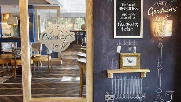Een restaurantinterieur met een prikbord waarop een gastvrije boodschap staat, een ingelijst beeld en een bordje met de tekst 'Goodwin's Kitchen'.