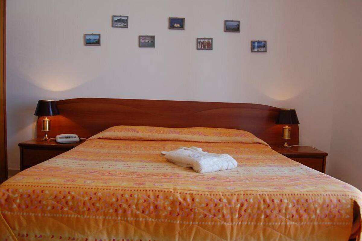 Un lit bien fait, recouvert d’une couvre-lit à motifs, deux lampes de chevet, un téléphone et un mur orné de plusieurs petits tableaux encadrés.
