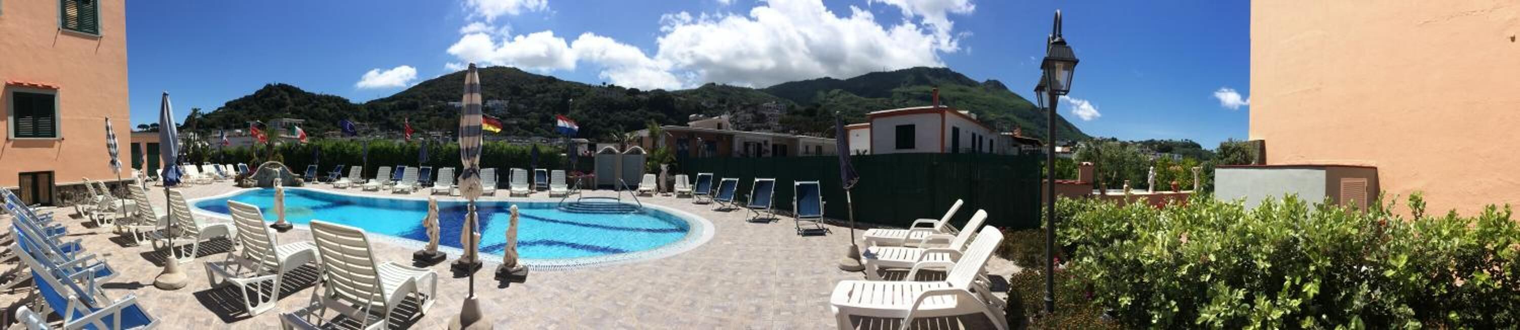A pool area with lounge chairs and umbrellas, surrounded by buildings and mountains.