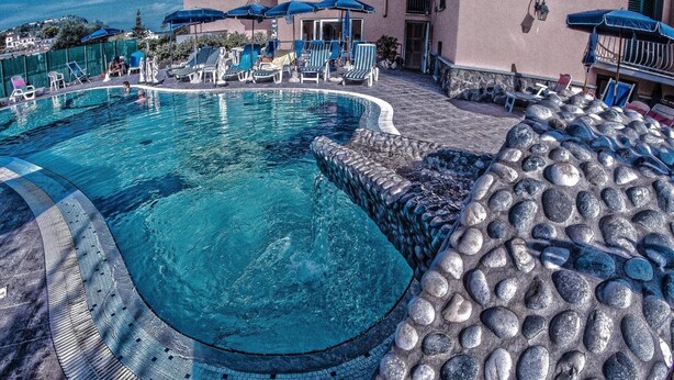 Ein Pool mit Steinrand, Liegestühlen und Sonnenschirmen.