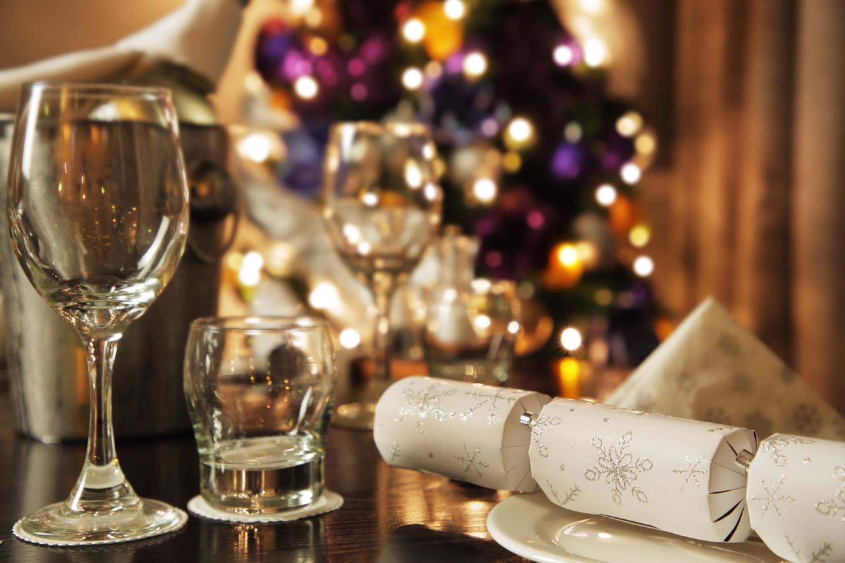 Une table avec des verres à vin et un menu enroulé, placée sur un fond légèrement éclairé avec un sapin de Noël flou en arrière-plan.
