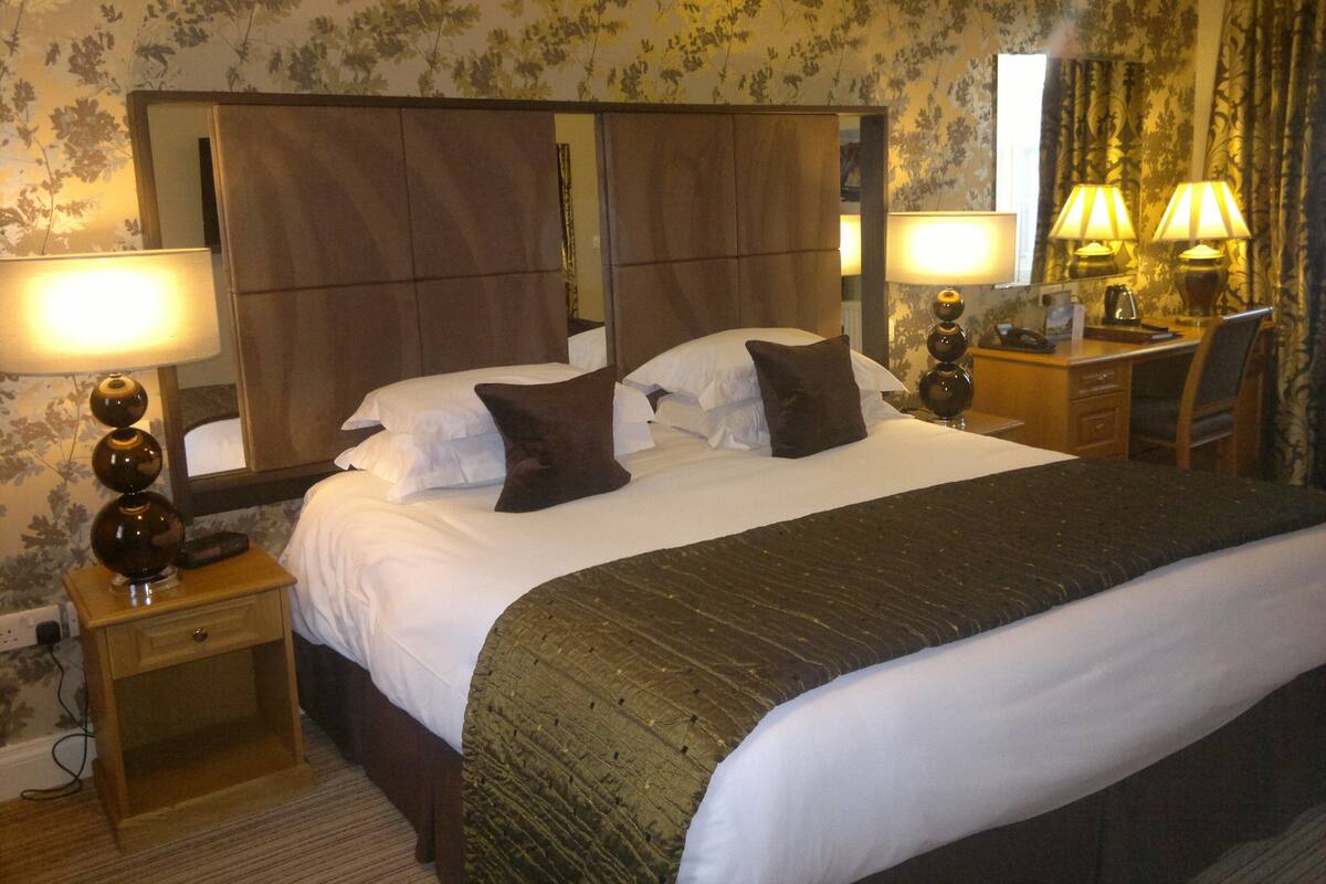 A hotel room with a large bed, two bedside tables with lamps, a desk with a chair, and a large mirror above the bed.