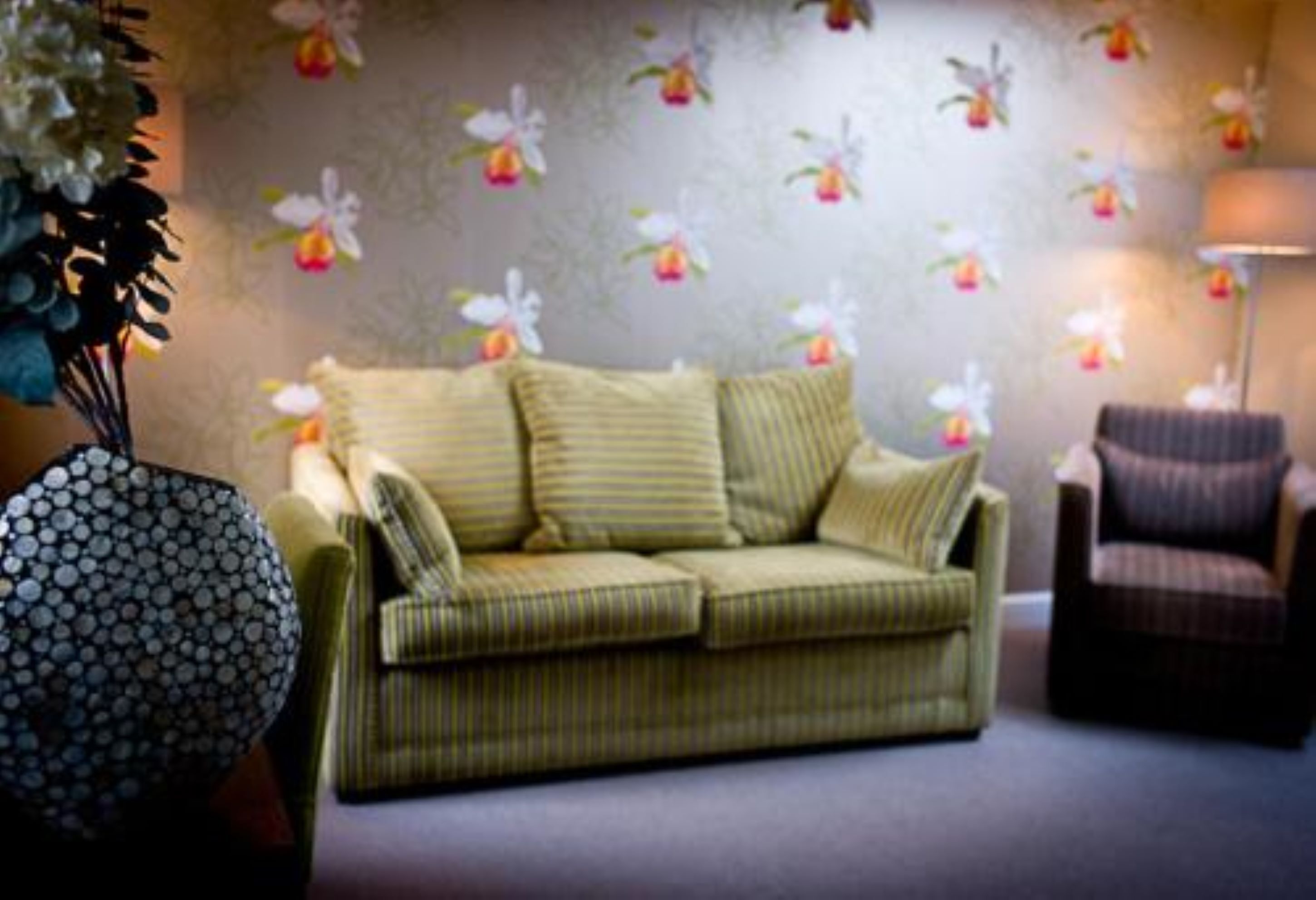 A living room with a green sofa, striped cushions, a patterned armchair, and a wall with floral wallpaper.