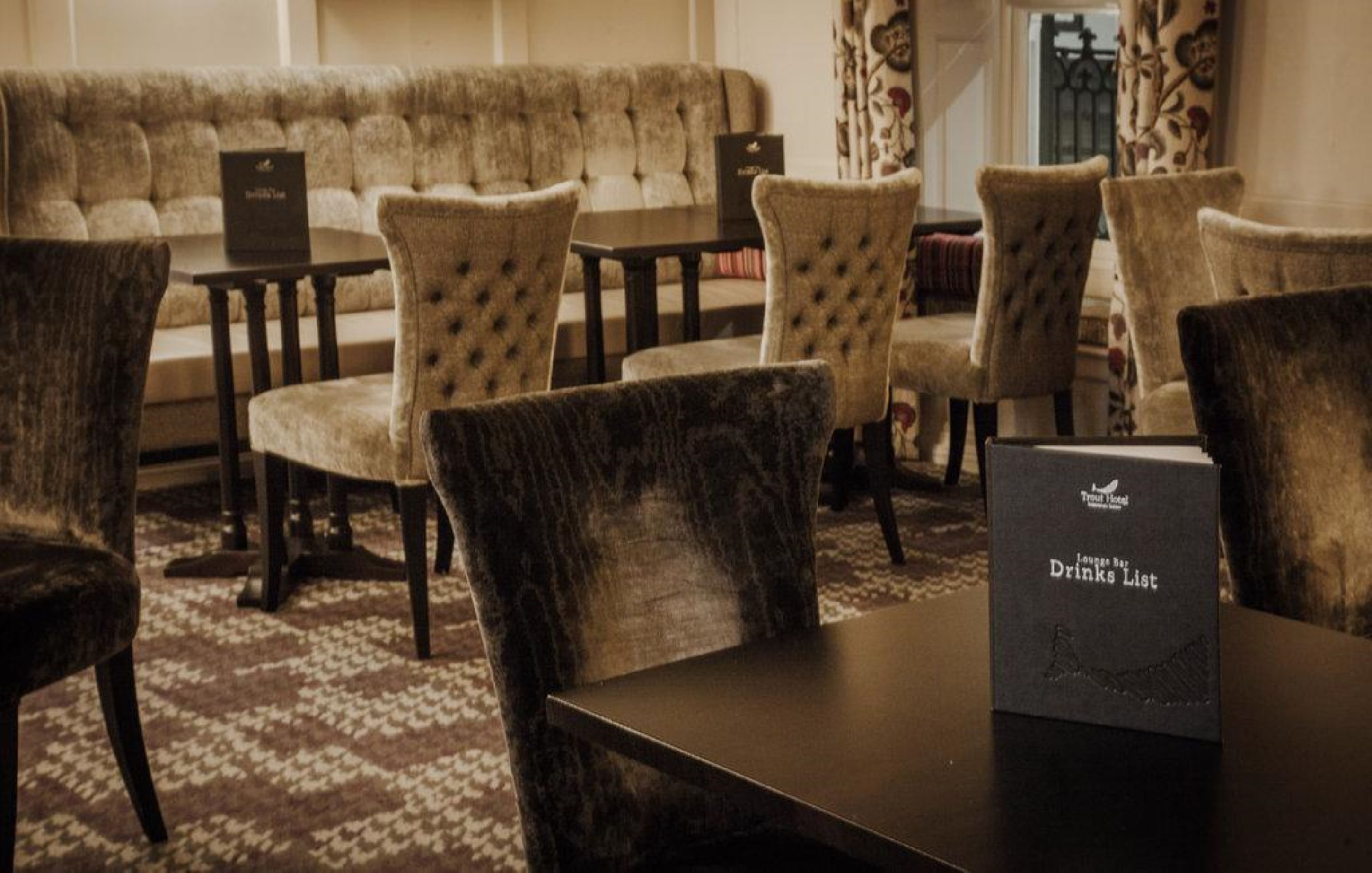 Un área de descanso con un sofá y varios sillones, una mesa con un expositor de la carta de bebidas, y una alfombra estampada.