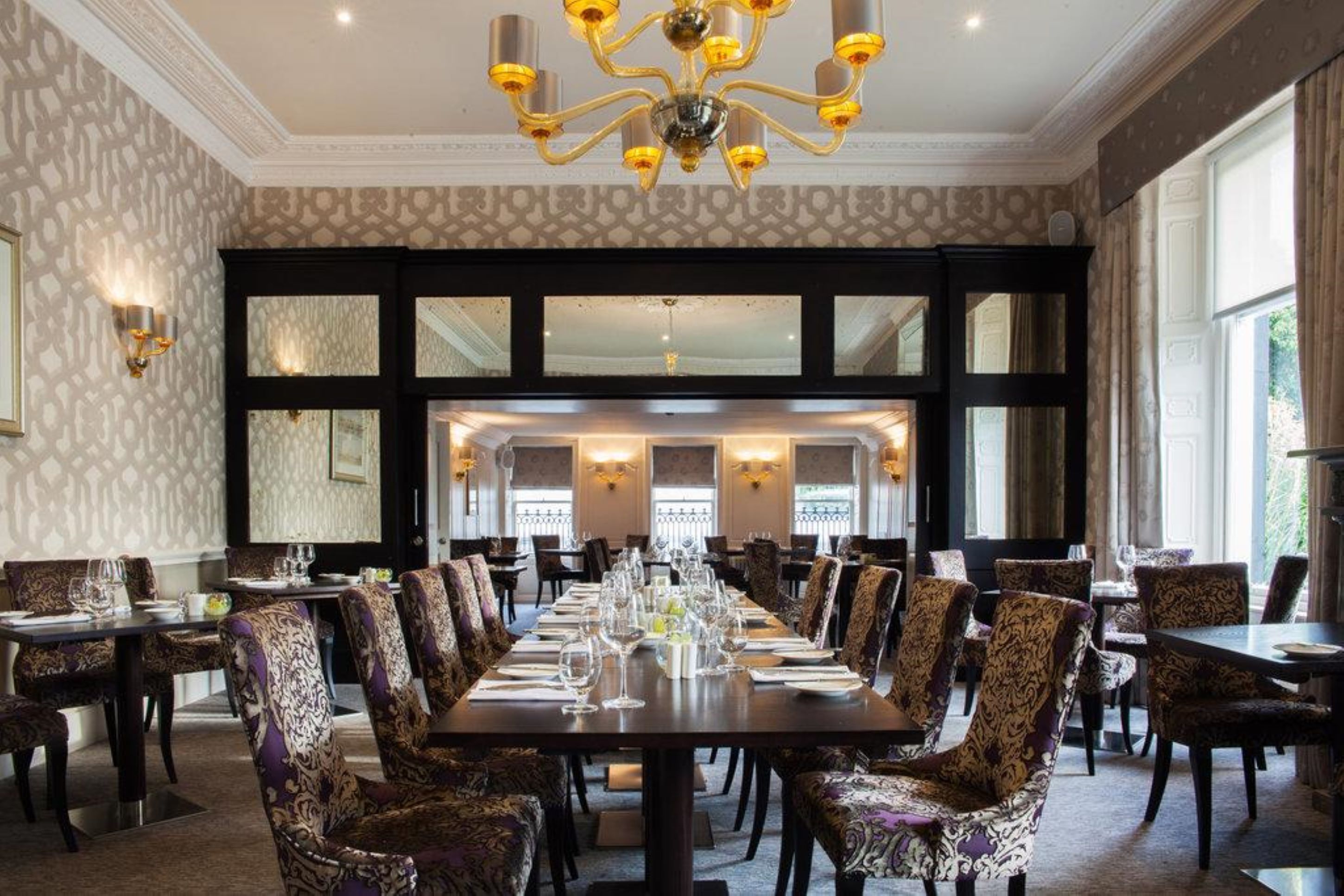 A formal dining room with a long table set for a meal, surrounded by patterned chairs, and a chandelier above.