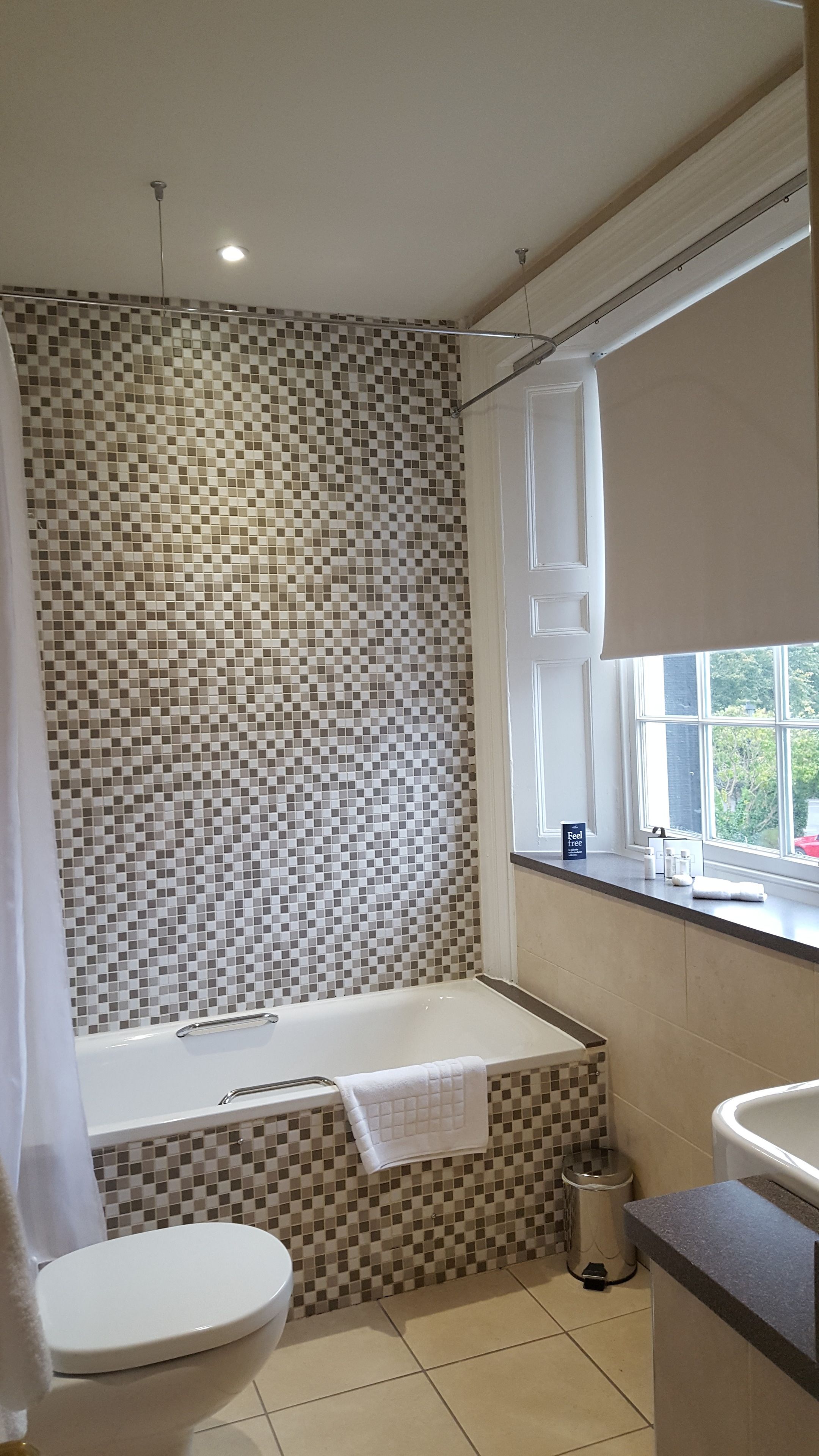 A bathroom with a mosaic tile wall, a bathtub, a toilet, and a window with a view of the outside.