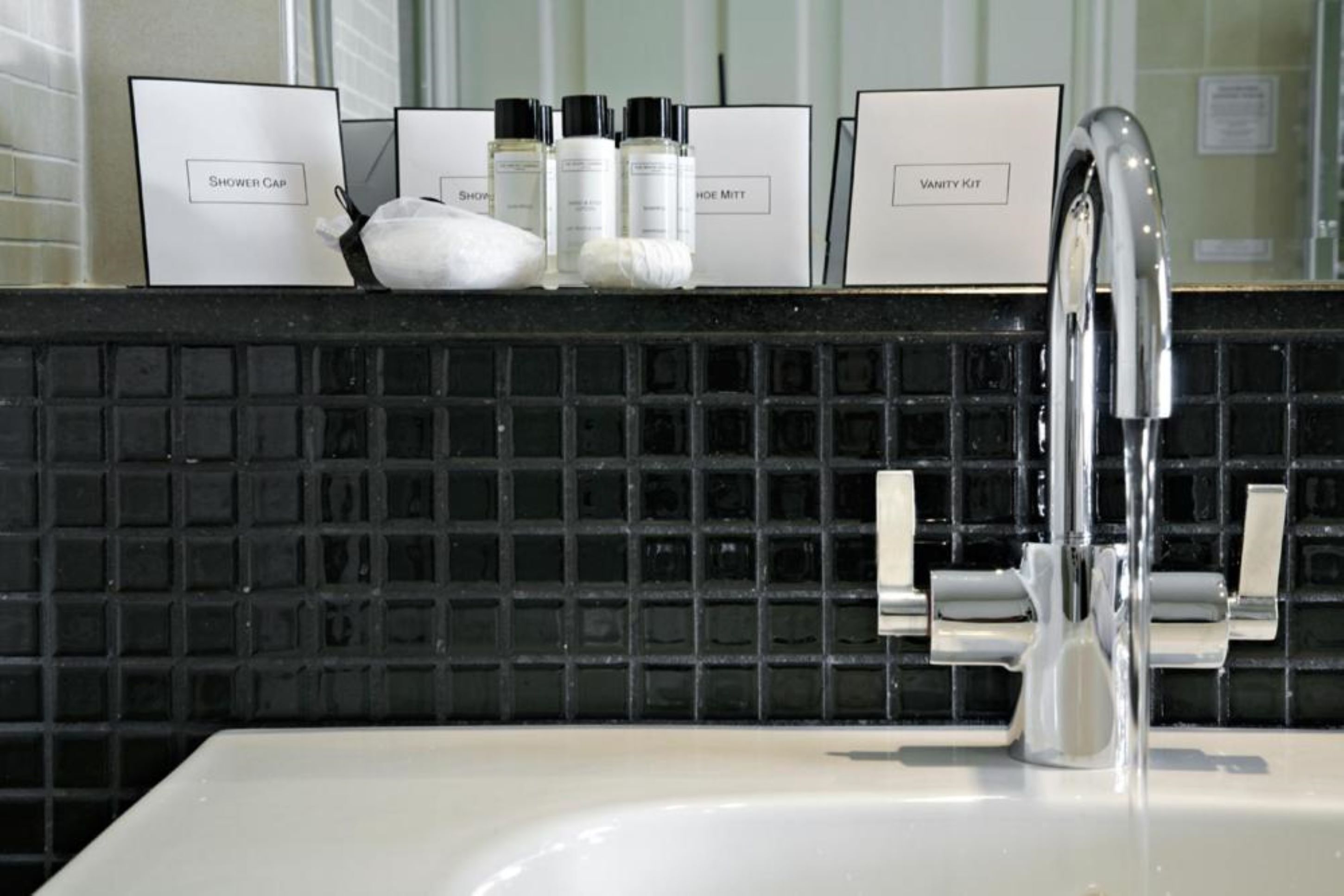 A bathroom with a black tiled backsplash, a white sink, and a vanity area with labeled storage boxes for shower cap, toiletries, and vanity kit.
