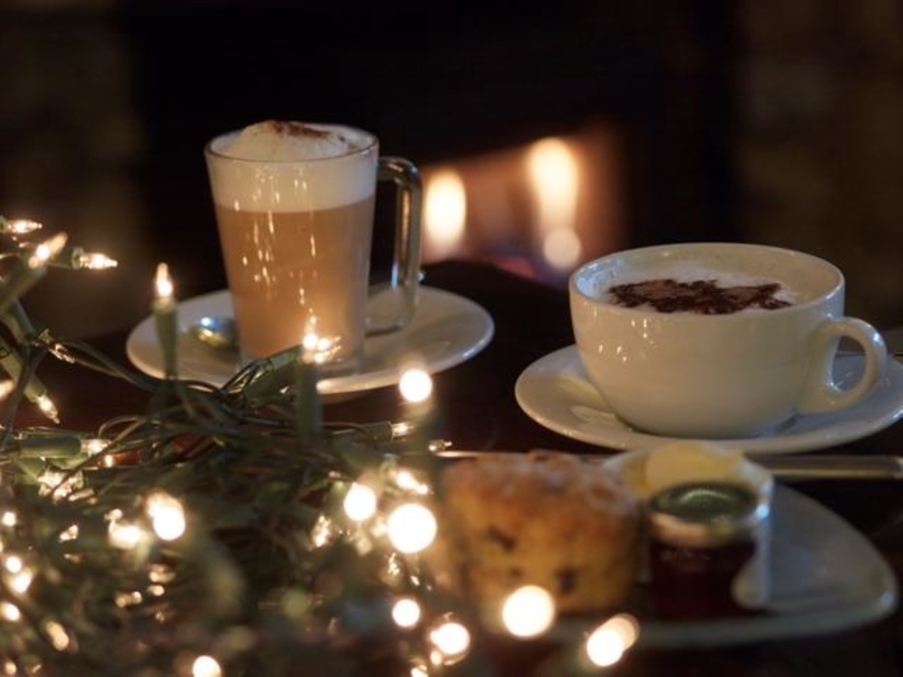 Dos tazas de café sobre platillos, un pastel pequeño y una guirnalda de luces en un ambiente tenue.